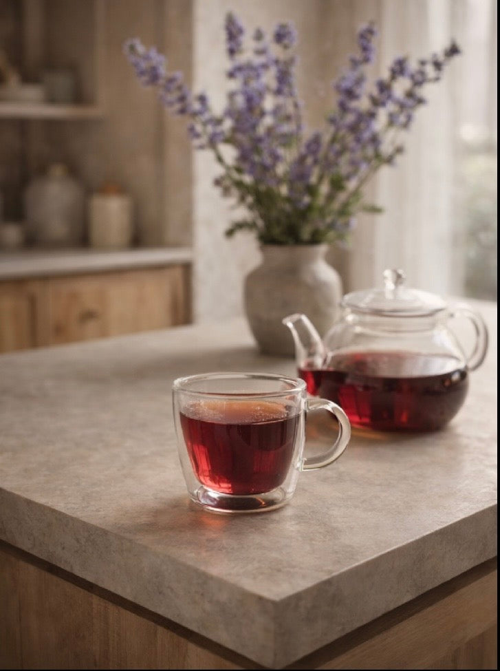 Glass teacup and teapot filled with a dark liquid on a kitchen counter with a vase of lavender in the background.